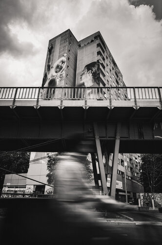 A person is riding a bicycle and a building is in the background with a train track. Long exposure street view in Germany. by ihsan Okur, 사진 for Sale on Singulart